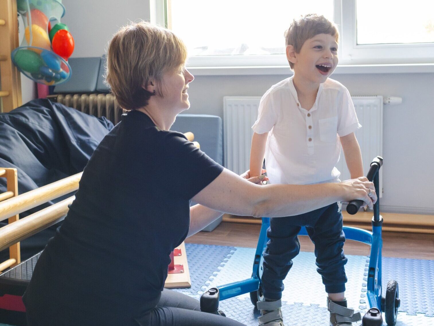 A woman assists a child in a room, with a walker positioned nearby, highlighting their supportive interaction.